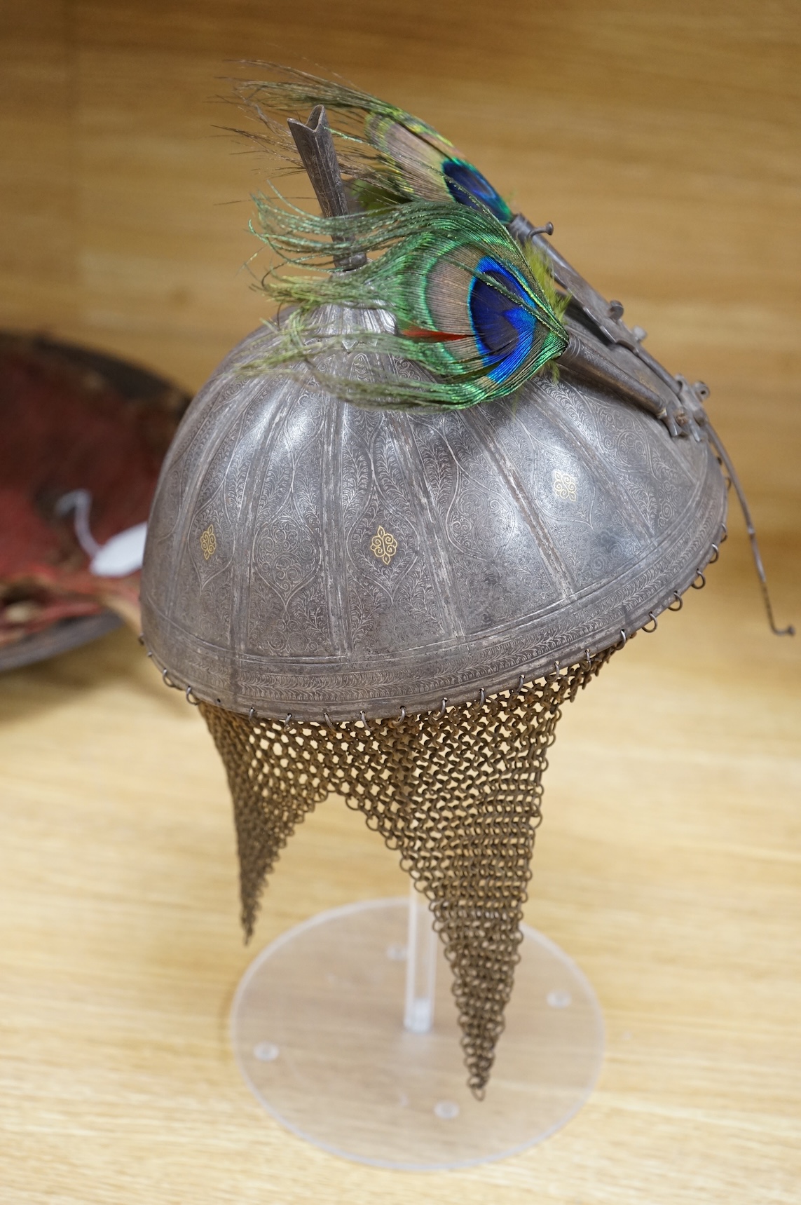A mid to late 19th century Indian helmet and matching shield, covered with silver koftgari decoration, the helmet with sliding nasal bar and twin plume sockets surmounted by a third plume socket, the skull fluted like a